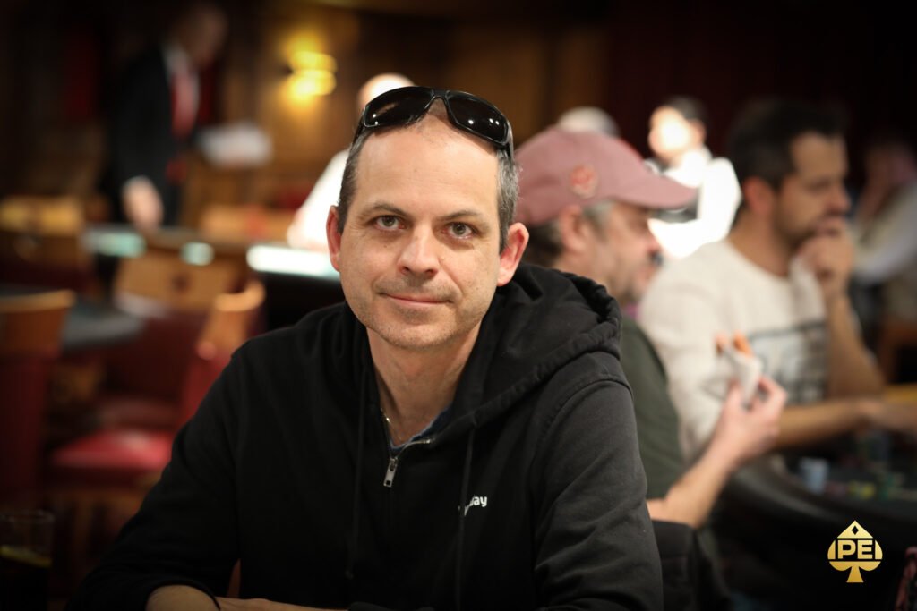 Ein Mann sitzt an einem Pokertisch und hält zwei Spielkarten in der Hand. Die Karten sind mit dem Bild eines Totenkopfes und gekreuzten Knochen bedruckt. Der Mann trägt