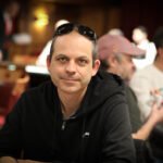 Ein Mann sitzt an einem Pokertisch und hält zwei Spielkarten in der Hand. Die Karten sind mit dem Bild eines Totenkopfes und gekreuzten Knochen bedruckt. Der Mann trägt
