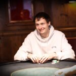 Ein Mann, der an einem Pokertisch sitzt, Karten hält und konzentriert auf seine Hand schaut, umgeben von anderen Spielern in einem Casino.