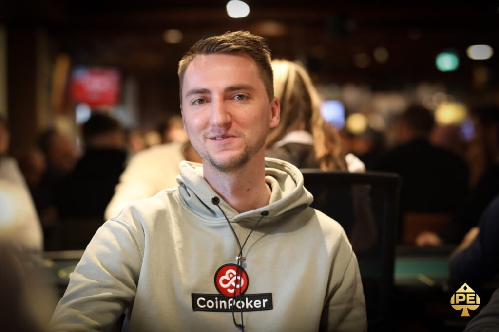 Ein Mann sitzt an einem Pokertisch und hält zwei Spielkarten in der Hand. Er schaut konzentriert auf den Tisch, umgeben von Pokerchips und anderen Spielern im Hintergrund.