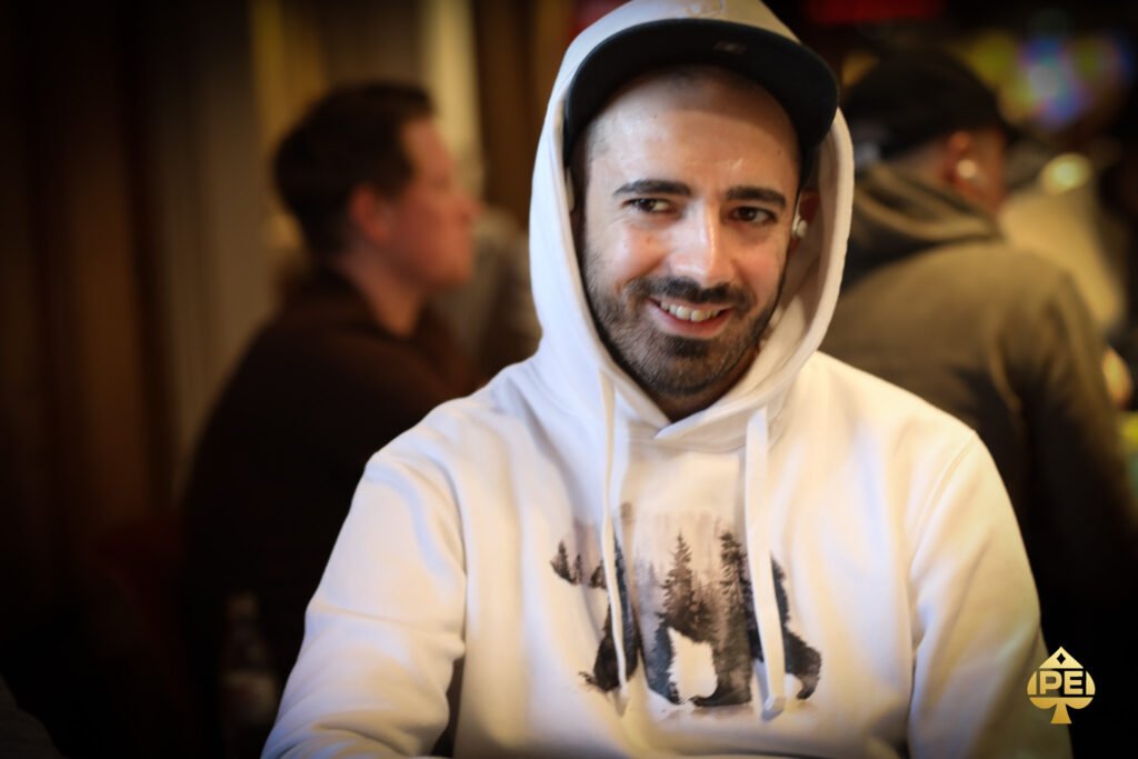 Ein Mann in einem schwarzen Anzug und einer Krawatte sitzt an einem Pokertisch mit Pokerchips und Karten. Er hat eine ernste Miene und schaut konzentriert auf seine Karten. Im Hinter