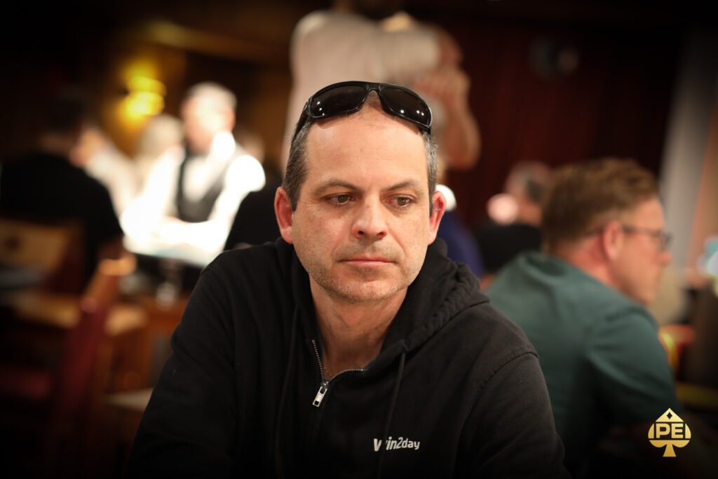 Ein Mann in einem dunklen Anzug und einer roten Krawatte spielt entschlossen Poker mit ernstem Gesichtsausdruck an einem eleganten Tisch mit grünem Filz und Chips.