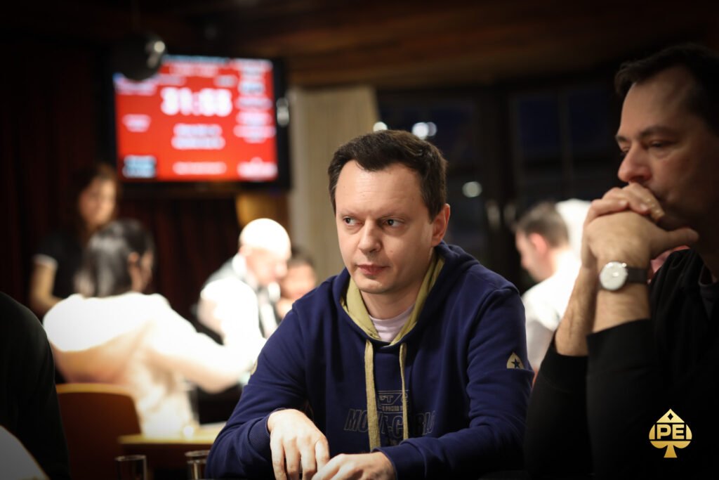 Ein Mann in einem schwarzen Anzug und einer schwarzen Sonnenbrille sitzt an einem Pokertisch und hält zwei Spielkarten in der Hand. Er wirkt konzentriert und ernsthaft, während er auf seine