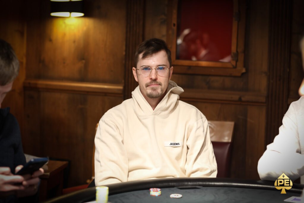 Ein Mann in einem schwarzen Anzug und einer Sonnenbrille sitzt an einem Pokertisch und hält zwei Karten in der Hand. Er schaut konzentriert auf seine Karten, während im Hintergrund andere Spieler
