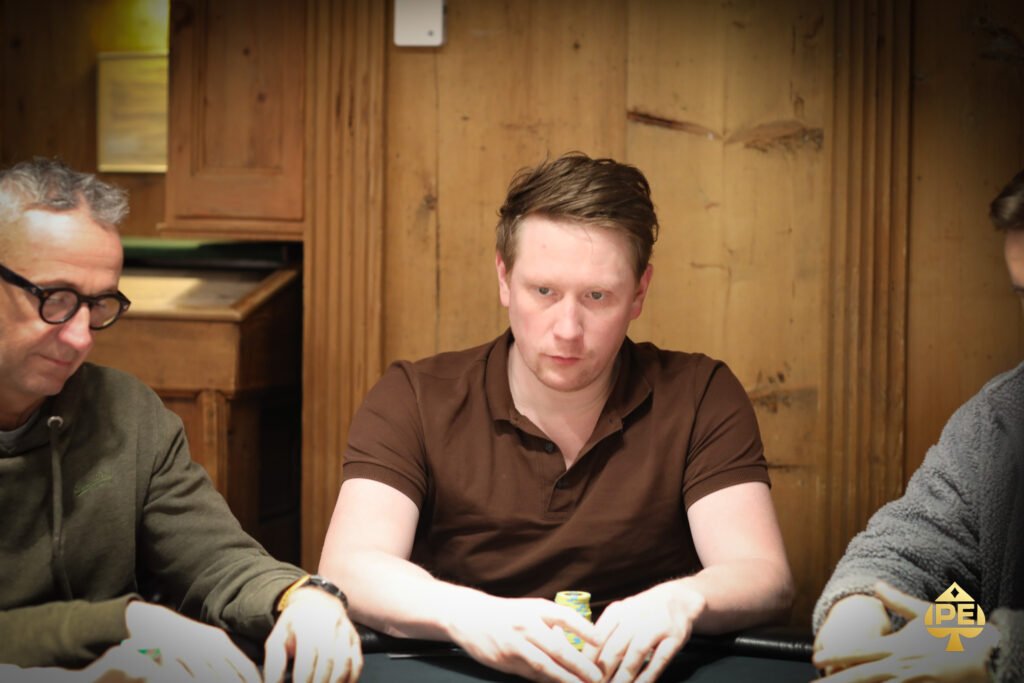 Ein Mann in einem schwarzen Anzug sitzt an einem Pokertisch und hält zwei Spielkarten in der Hand. Er schaut konzentriert auf die Karten. Im Hintergrund sind Pokerchips und andere Spieler zu