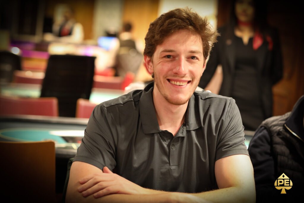 Ein Mann in einem Anzug sitzt an einem Pokertisch und zeigt eine Siegerhand mit einem breiten Lächeln. Andere Spieler sitzen um den Tisch herum und schauen gespannt zu.