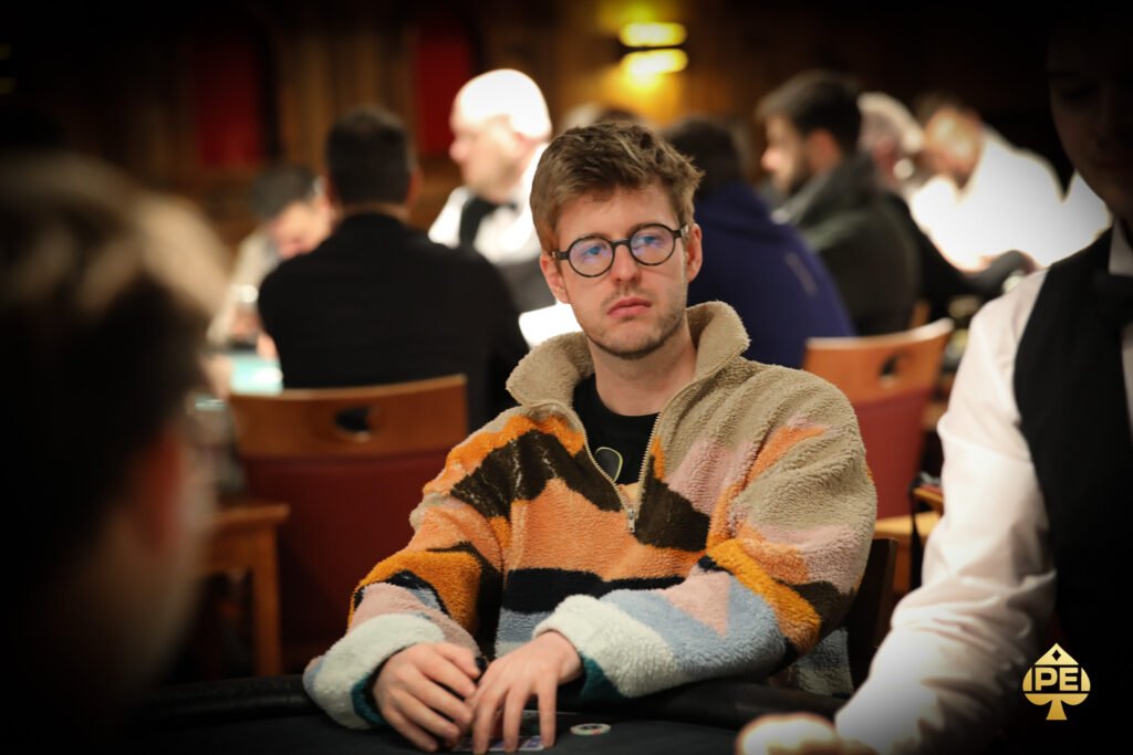 Ein Mann sitzt an einem Pokertisch mit Chips und Karten vor sich. Er hat eine konzentrierte Miene und trägt eine schwarze Kappe.