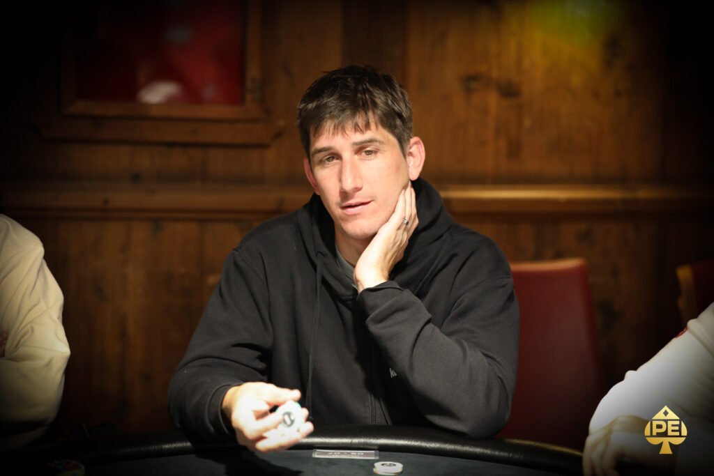 Ein Mann in einem schwarzen Anzug und einer roten Krawatte sitzt an einem Pokertisch mit grüner Filzdecke. Er hält seine Karten in der Hand und schaut konzentriert auf den T