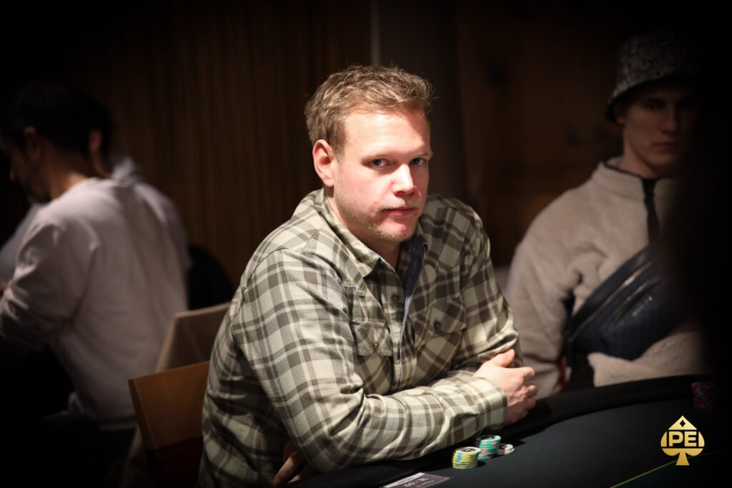 Ein Mann und eine Frau sitzen an einem Pokertisch und spielen eine Partie Poker. Der Mann hat eine Hand mit Karten in der Hand, während die Frau konzentriert auf ihre eigenen Karten schaut. Im Hintergrund