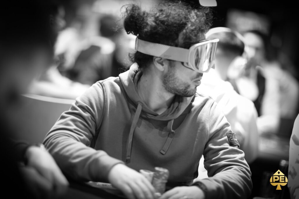 Ein Mann sitzt an einem Pokertisch und hält zwei Karten in der Hand, während er konzentriert auf die Gemeinschaftskarten in der Mitte des Tisches schaut. Andere Spieler und Dealer sind im H