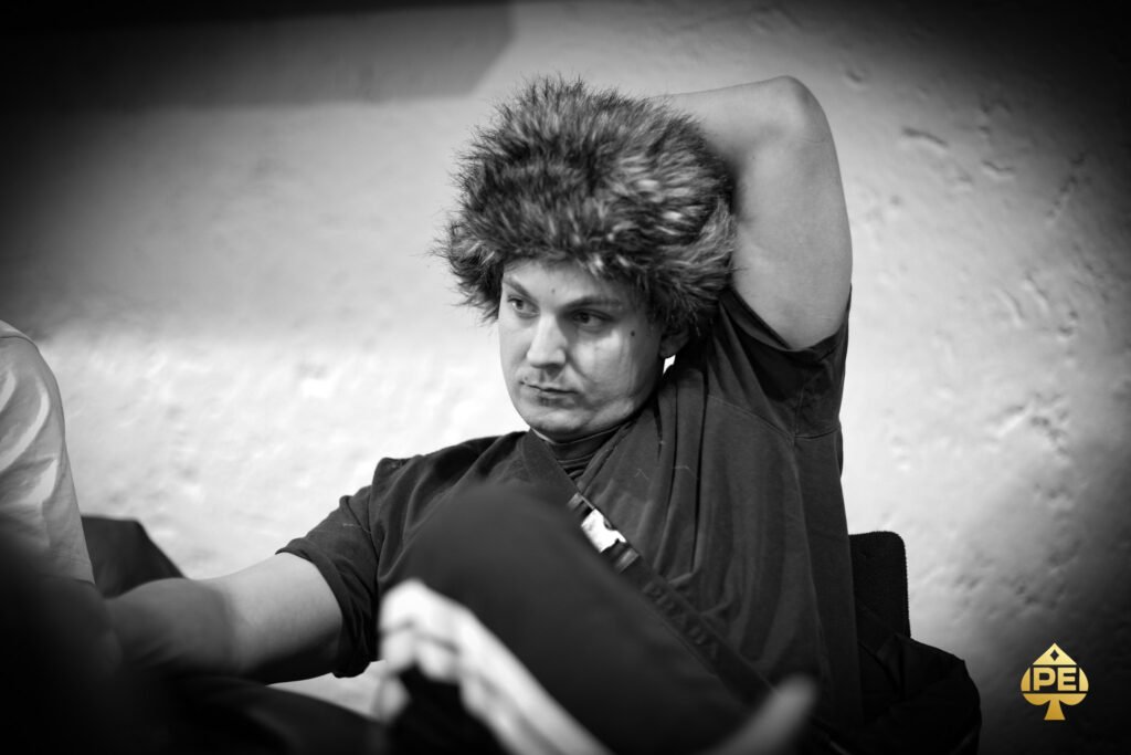 Ein Mann und eine Frau sitzen an einem Pokertisch und betrachten konzentriert ihre Karten. In der Mitte des Tisches liegen Pokerchips und Spielkarten. Im Hintergrund sind weitere Spieler und eine bele