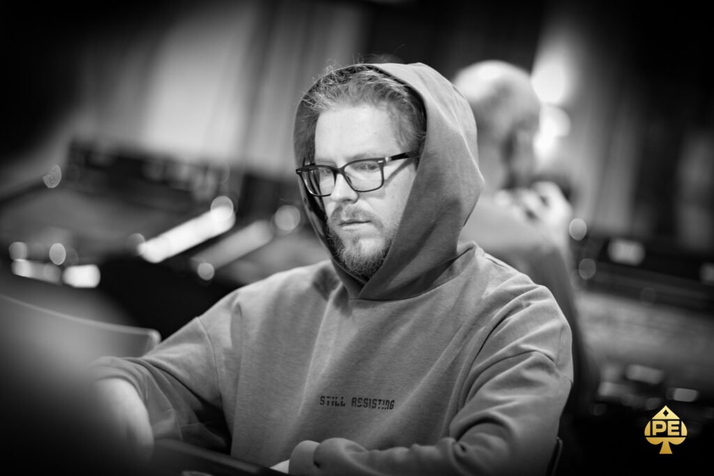 Ein Mann sitzt an einem Pokertisch und hält zwei Karten in der Hand, während er konzentriert auf die Gemeinschaftskarten schaut. Um ihn herum sind Pokerchips und ein Stapel Karten zu