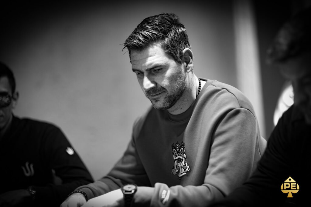Ein Mann in einem dunkelblauen Anzug und einer blauen Krawatte steht vor einem Pokertisch mit Chips und Karten. Er hält zwei Spielkarten in der Hand und schaut konzentriert auf den Tisch
