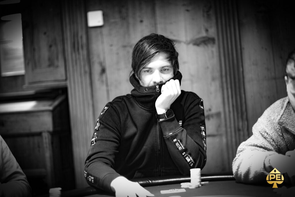 Ein Mann in einem schwarzen Anzug und einer schwarzen Sonnenbrille sitzt an einem Pokertisch und hält seine Karten. Er wirkt konzentriert und selbstbewusst. Im Hintergrund sind andere Pok