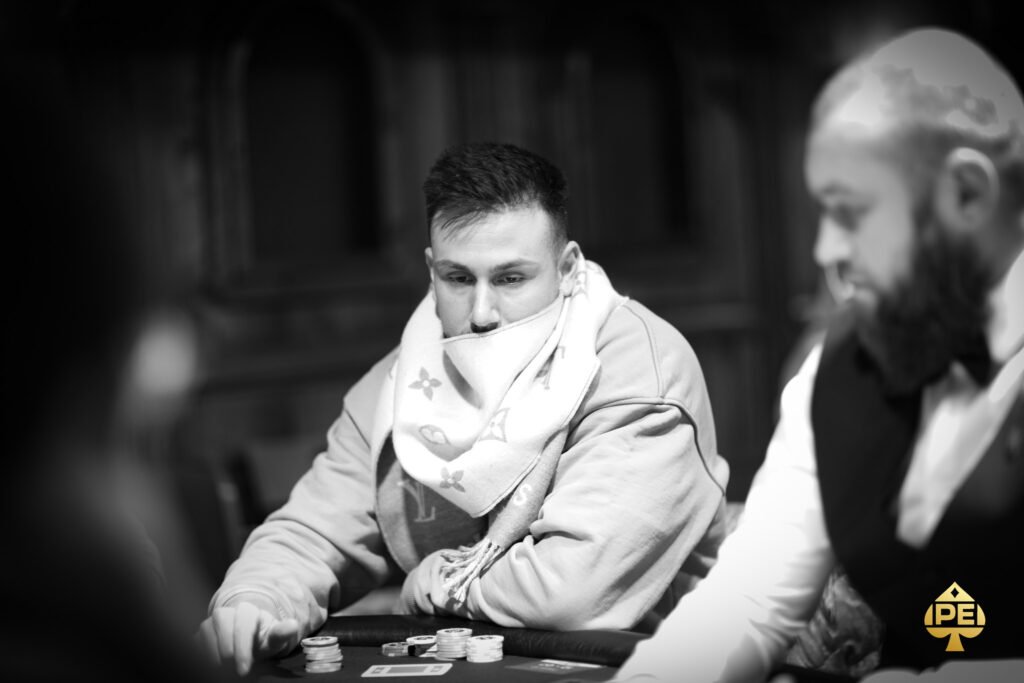 Ein Mann in einem dunkelblauen Anzug und einer hellblauen Krawatte sitzt an einem Pokertisch mit grünem Filz. Er hält Karten in der Hand und schaut konzentriert auf den T