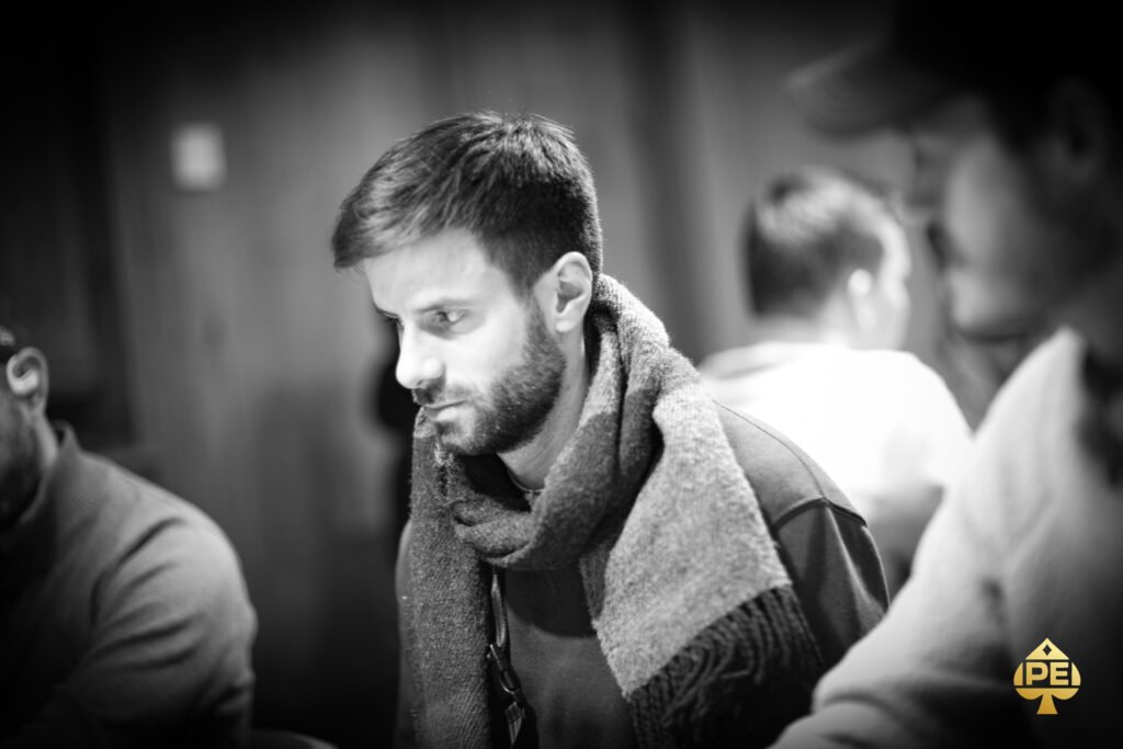 Ein Mann sitzt an einem Pokertisch und betrachtet konzentriert seine Karten. Um ihn herum sind weitere Spieler zu sehen, die ebenfalls ihre Karten prüfen. Im Hintergrund sind Pokerchips und Get