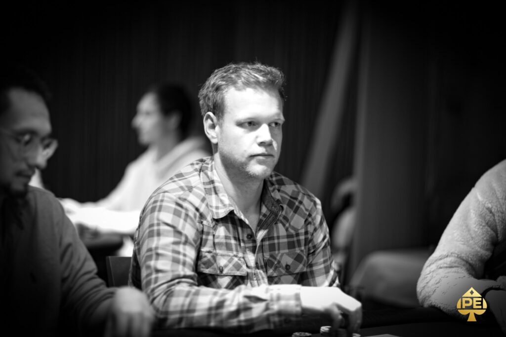 Ein Mann sitzt an einem Pokertisch und hält zwei Spielkarten in der Hand. Er hat eine konzentrierte Miene und schaut auf seine Karten. Im Hintergrund sind Chips und Spielkarten auf dem T