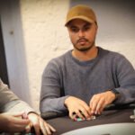 Ein Mann sitzt an einem Pokertisch und hält seine Karten in der Hand. Er wirkt konzentriert und fokussiert. Um ihn herum sind Pokerchips und andere Spieler zu sehen.