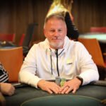Ein Mann und eine Frau sitzen am Pokertisch und halten ihre Karten. Sie haben konzentrierte Gesichtsausdrücke. Um sie herum sind Pokerchips und Getränke zu sehen.