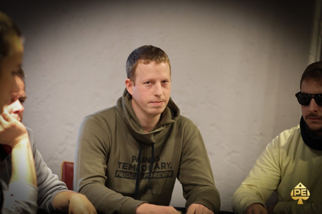 Ein Mann in einem schwarzen Anzug sitzt an einem Pokertisch und hält zwei Spielkarten in der Hand. Er scheint konzentriert und selbstbewusst zu sein, während er an einem Pokerspiel teilnim