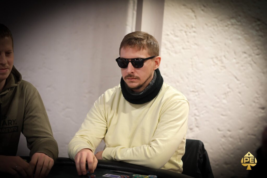 Ein Mann in einem schwarzen Anzug und einer schwarzen Sonnenbrille spielt Poker an einem eleganten Tisch in einem Casino, umgeben von anderen Spielern und einer Dealerin.