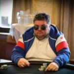 Ein Mann und eine Frau sitzen an einem Pokertisch und halten Karten in der Hand. Sie schauen konzentriert auf ihre Karten und setzen Pokerchips. Im Hintergrund sind andere Spieler und ein Dealer zu sehen.