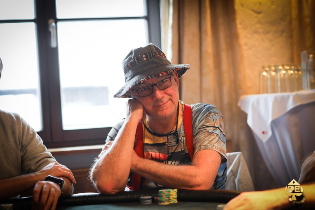 Ein Mann und eine Frau sitzen an einem Pokertisch und spielen eine Partie Poker. Der Mann hält Karten in der Hand, während die Frau konzentriert auf ihre Chips schaut. Im Hintergrund sind weitere Spieler und