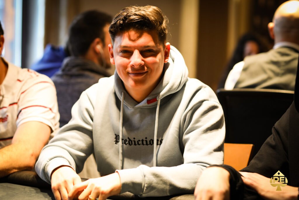 Ein Mann in einem dunkelblauen Anzug sitzt an einem Pokertisch und hält eine Hand mit Spielkarten. Er wirkt konzentriert und fokussiert. Die anderen Spieler am Tisch sind unscharf