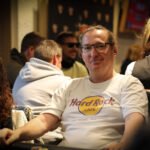 Ein professioneller Pokertisch mit grüner Filzoberfläche, umgeben von Spielkarten, Pokerchips und Getränken. Mehrere Spieler sitzen um den Tisch herum und konzentrieren sich auf