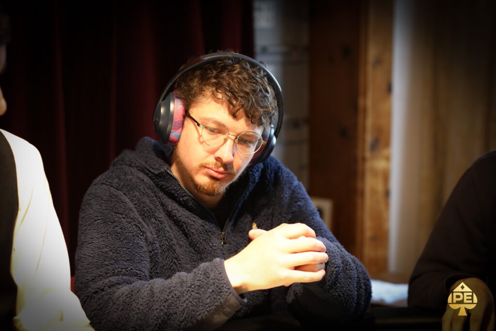 Ein Mann in einem schwarzen Anzug sitzt an einem Pokertisch und hält Karten in der Hand. Im Hintergrund sind andere Spieler und Pokerchips zu sehen.