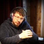 Ein Mann in einem schwarzen Anzug sitzt an einem Pokertisch und hält Karten in der Hand. Im Hintergrund sind andere Spieler und Pokerchips zu sehen.
