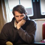Ein Mann in einem dunklen Anzug sitzt an einem Pokertisch mit grüner Filzoberfläche. Er hält Karten in der Hand und schaut konzentriert auf den Tisch. Im Hintergrund sind