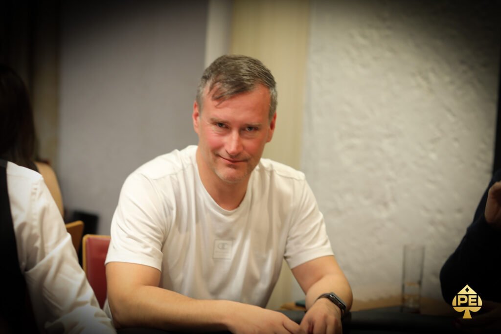 Ein Mann in einem schwarzen Anzug und einer roten Krawatte sitzt an einem Pokertisch und hält einen Stapel Pokerchips in der Hand. Im Hintergrund sind weitere Spieler und grüne Filz-Tisch