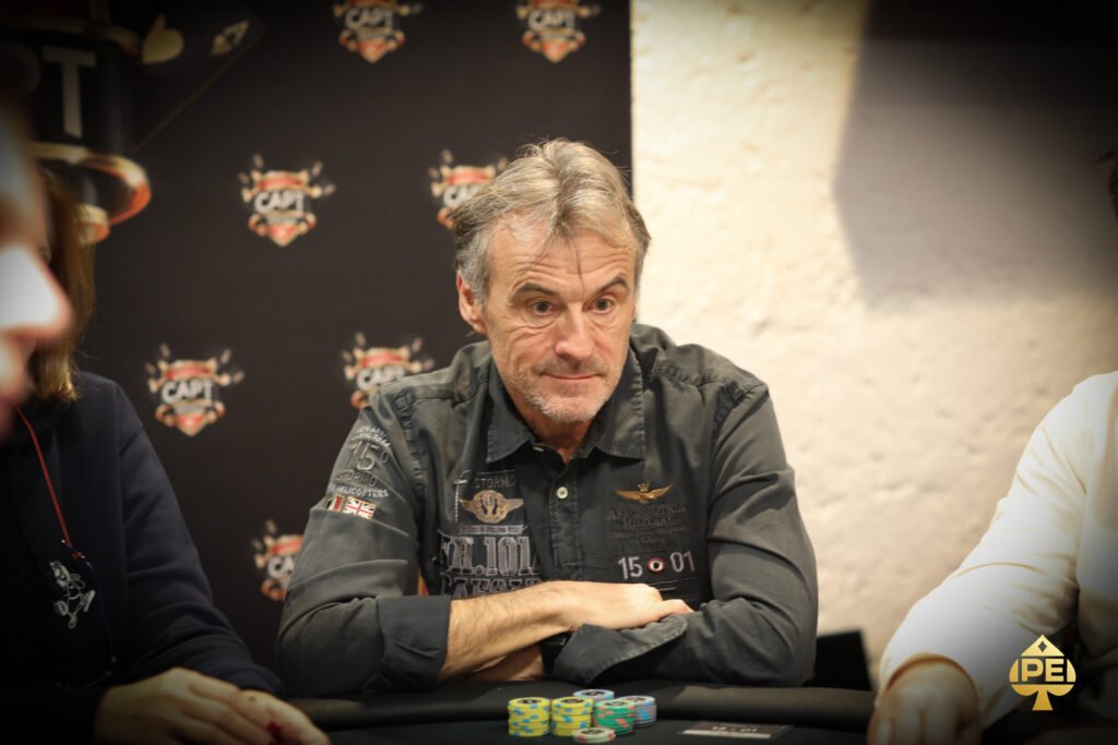 Ein professioneller Pokerspieler mit Sonnenbrille und Kappe sitzt an einem Tisch und hält seine Spielkarten. In der Hand hat er Pokerchips und im Hintergrund sind andere Spieler und Dealer zu sehen.