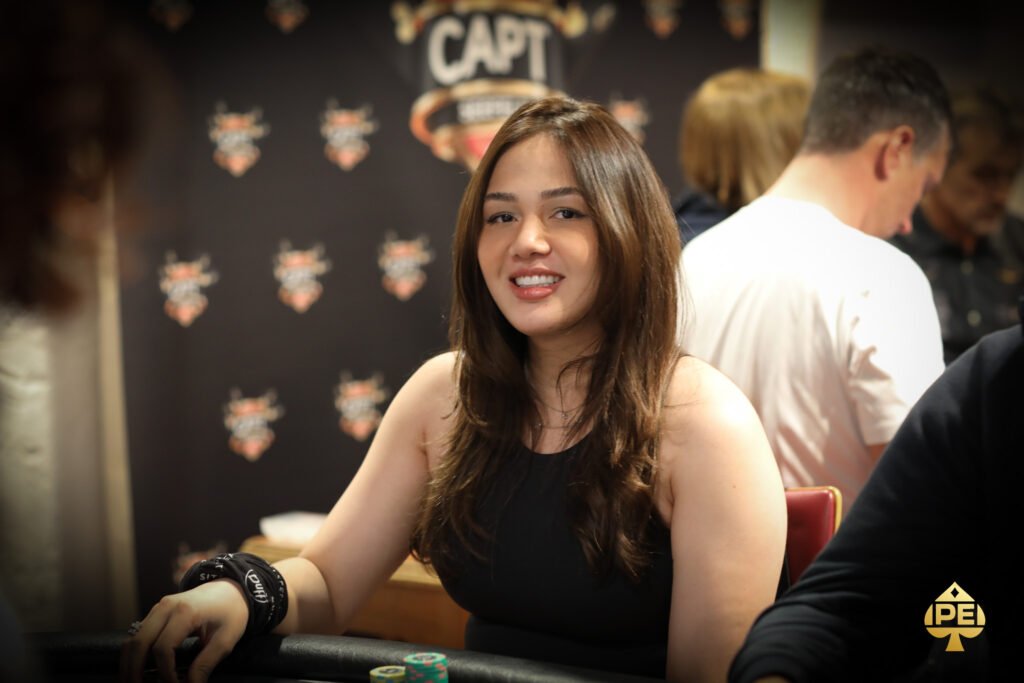 Ein professioneller Pokerspieler mit Sonnenbrille und Kappe sitzt an einem Pokertisch und betrachtet konzentriert seine Karten. Im Hintergrund sind Chips und Spielkarten zu sehen.