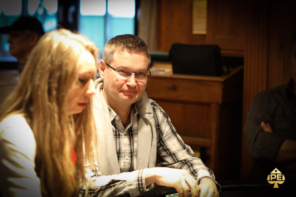 Ein Mann sitzt an einem Pokertisch und hält zwei Spielkarten in der Hand, während er konzentriert auf seine Gegner schaut. Die Tischkarten und Pokerchips sind ebenfalls auf dem Tisch verteilt
