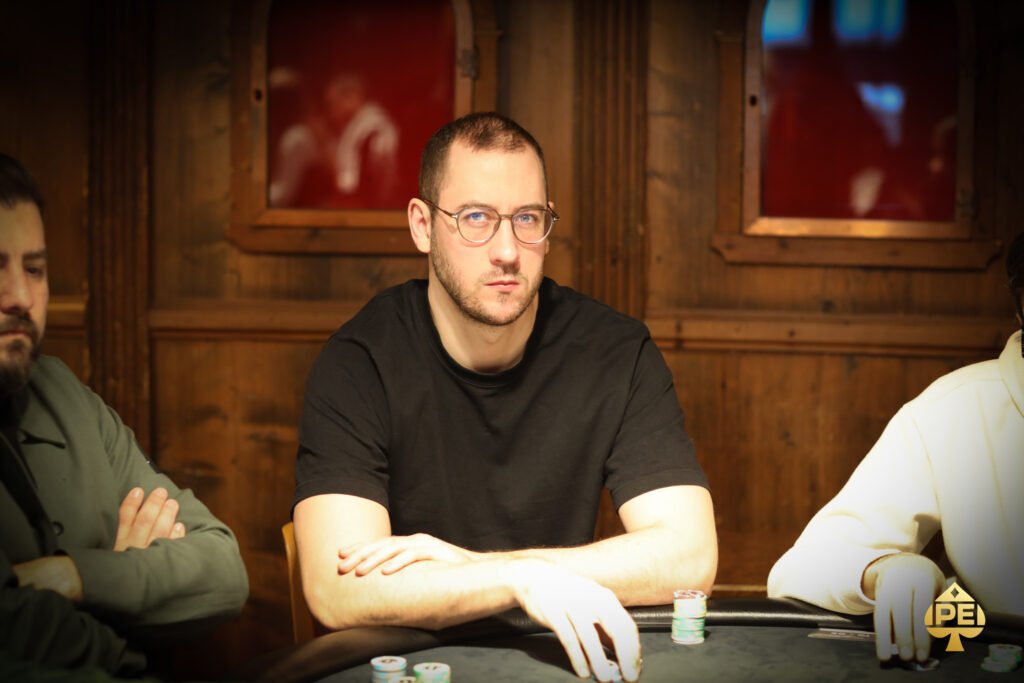 Ein Mann in einem Anzug und mit einer Sonnenbrille spielt Poker an einem Tisch mit Karten und Chips. Er hat eine ernste Miene und betrachtet konzentriert seine Karten. Andere Spieler sind im
