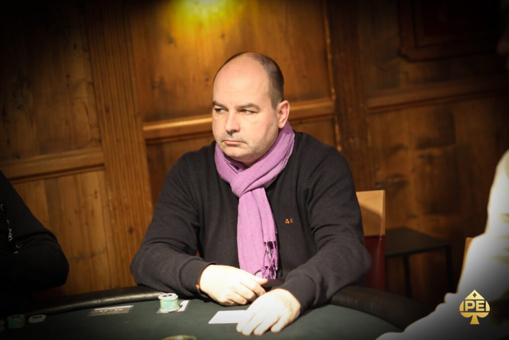 Ein professioneller Pokerspieler sitzt an einem Tisch mit Pokerchips und Karten, während er konzentriert in die Kamera schaut.