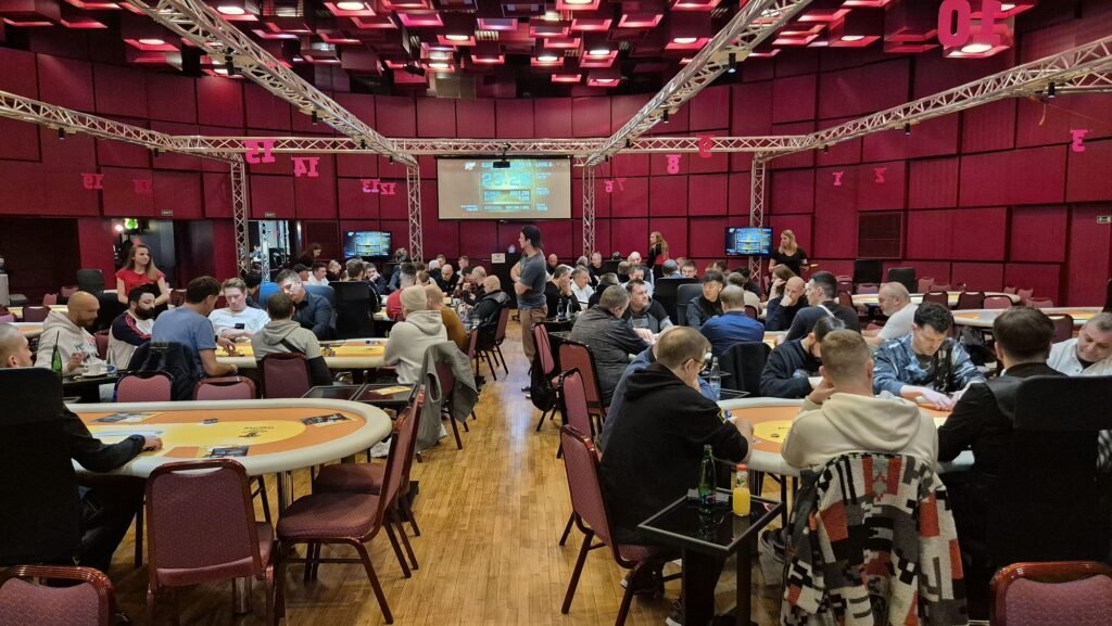 Ein Mann in einem dunkelblauen Anzug sitzt an einem Pokertisch und hält zwei Spielkarten in der Hand. Er schaut konzentriert auf den Tisch, auf dem Pokerchips liegen. Im H