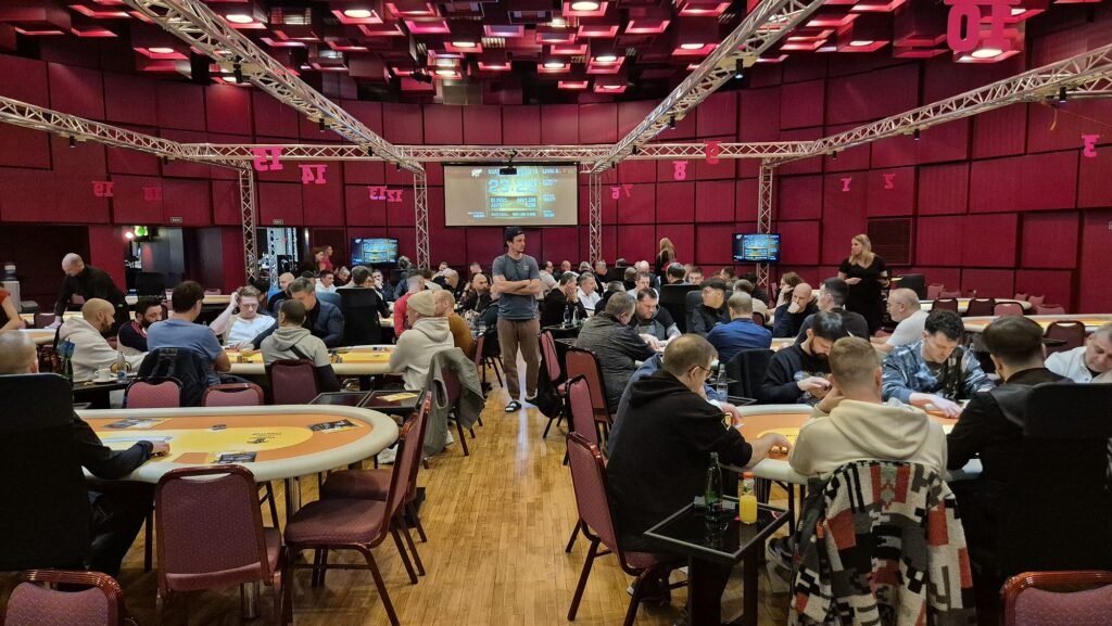 Ein Mann mit dunklen Haaren und Bart sitzt an einem Pokertisch und zeigt eine Hand mit einem König und einem Ass. Im Hintergrund sind Pokerchips und Karten zu sehen.