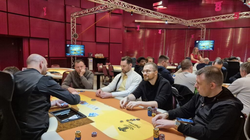 Ein Mann und eine Frau sitzen an einem Pokertisch und sehen sich konzentriert an, während sie Karten halten. Der Mann trägt ein schwarzes Hemd und die Frau ein rotes Kleid. Die Tischdecke