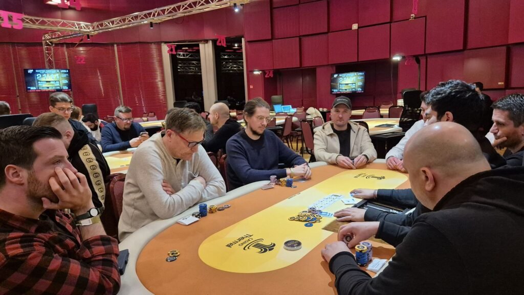 Ein Mann sitzt an einem Pokertisch und betrachtet konzentriert seine Karten. Um ihn herum stehen andere Spieler und beobachten gespannt das Spiel. Ein Stapel Pokerchips liegt in der Mitte des