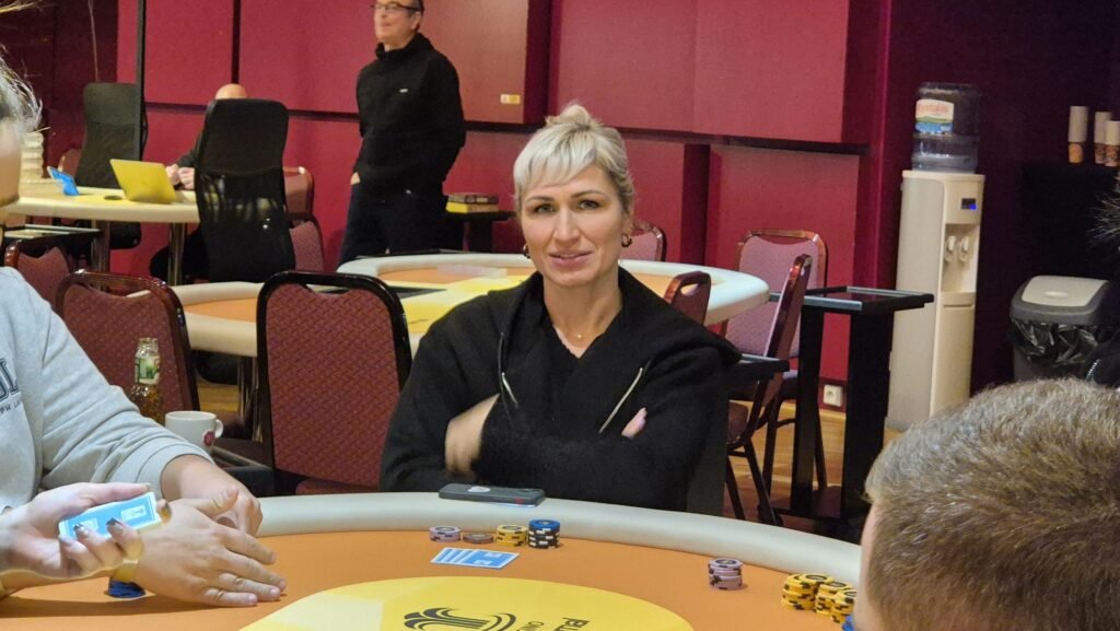 Ein Mann in einem eleganten Anzug sitzt vor einem Pokertisch, hält Karten in der Hand und schaut konzentriert auf den Tisch. Im Hintergrund sind andere Spieler und Pokerchips zu sehen.
