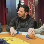 Ein Tisch mit einem grünen Pokertuch, auf dem Spielkarten und Pokerchips liegen. In der Mitte des Tisches befindet sich ein Stapel von Jetons. Im Hintergrund sind Spielerhände zu