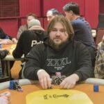 Ein Mann in einem dunkelblauen Anzug sitzt an einem Pokertisch mit grünem Filz. Er hält seine Spielkarten in der Hand und schaut konzentriert auf den Tisch. Im Hintergrund