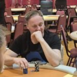 Ein Mann in einem dunklen Anzug und einer Krawatte sitzt an einem Pokertisch und hält zwei Spielkarten in der Hand. In der Mitte des Tisches liegen weitere Karten. Im Hintergrund sind andere