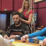 Ein Mann spielt Poker an einem Tisch mit grüner Filzdecke. Er hat eine Hand mit König-Karten. Andere Spieler und Pokerchips sind im Hintergrund zu sehen.