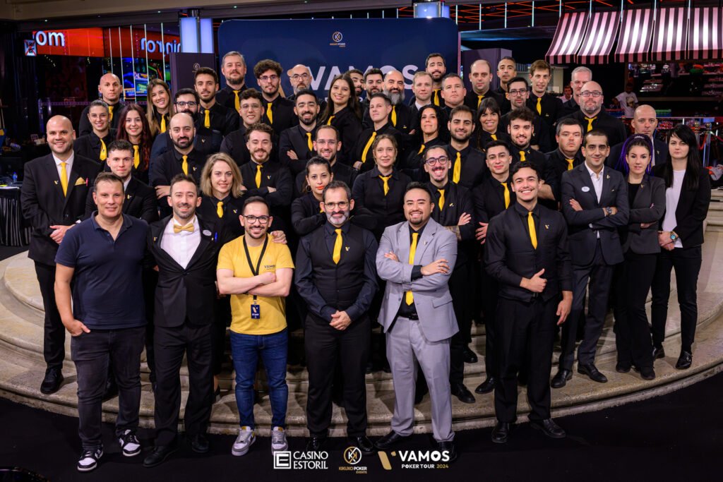 Ein Gruppenfoto von vier professionell gekleideten Pokerspielern, die lächelnd an einem Tisch sitzen und Karten halten. Einer der Spieler zeigt ein breites Siegerlächeln. Im Hinter
