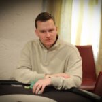 Ein Mann und eine Frau sitzen an einem Pokertisch und spielen Texas Hold'em. Der Mann hält Karten in der Hand, während die Frau mit ernstem Gesichtsausdruck auf ihre eigenen Karten schaut. Im H