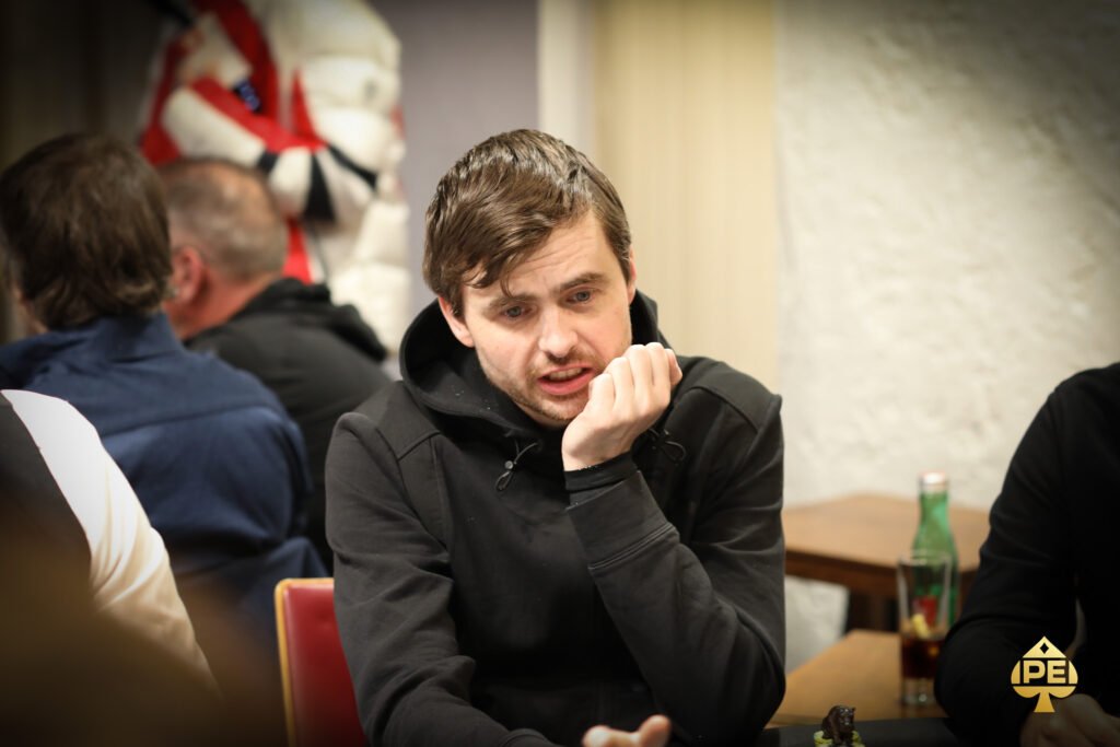 Ein Mann und eine Frau sitzen an einem Pokertisch und spielen Texas Hold'em. Der Mann hält Karten in der Hand und schaut konzentriert auf den Tisch, während die Frau mit gespielter Gelassenheit ihre
